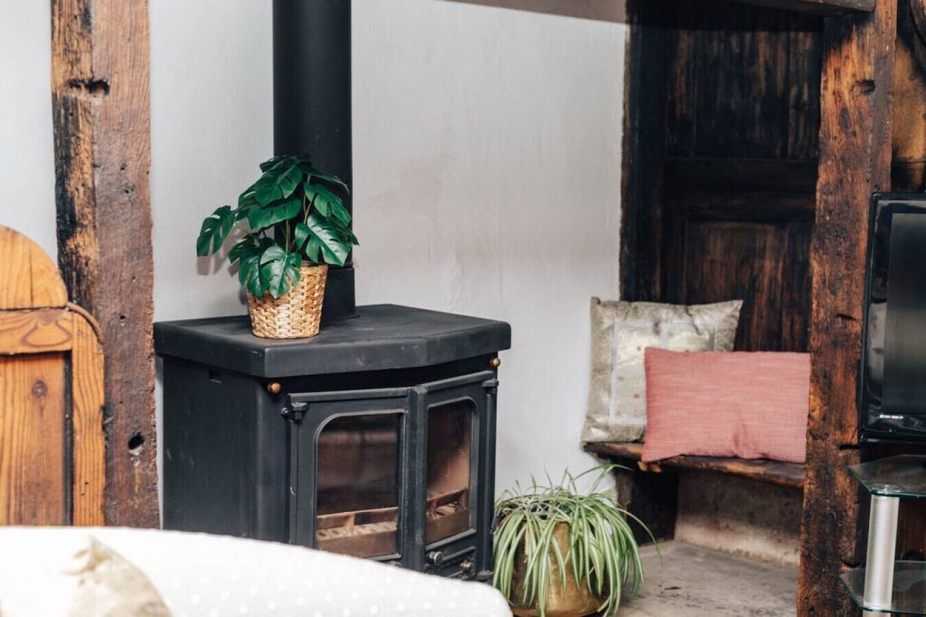 A cozy room with a black wood-burning stove, a wicker basket with a plant, a wooden door, and a bed with a pink pillow.