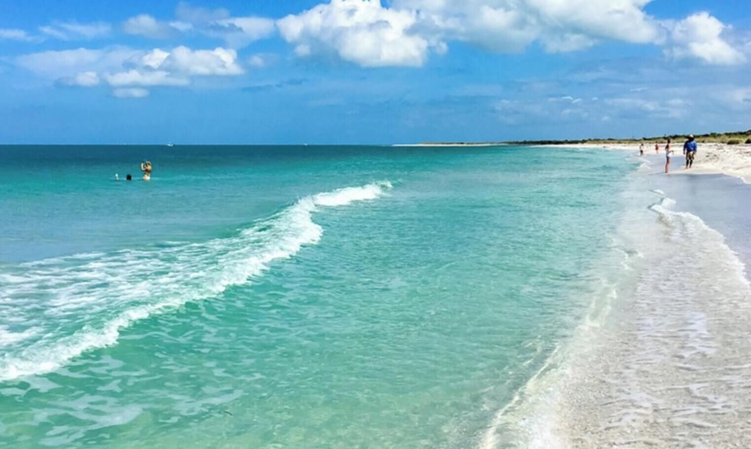 A beach with clear turquoise water, a sandy shore, and a few people walking.