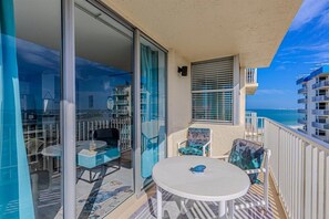 A balcony with a glass door, a round table, two chairs, and a view of the ocean and buildings. - Steps to the Sand, Dive into the Pool – Live the Dream!" (Fort Myers Beach)