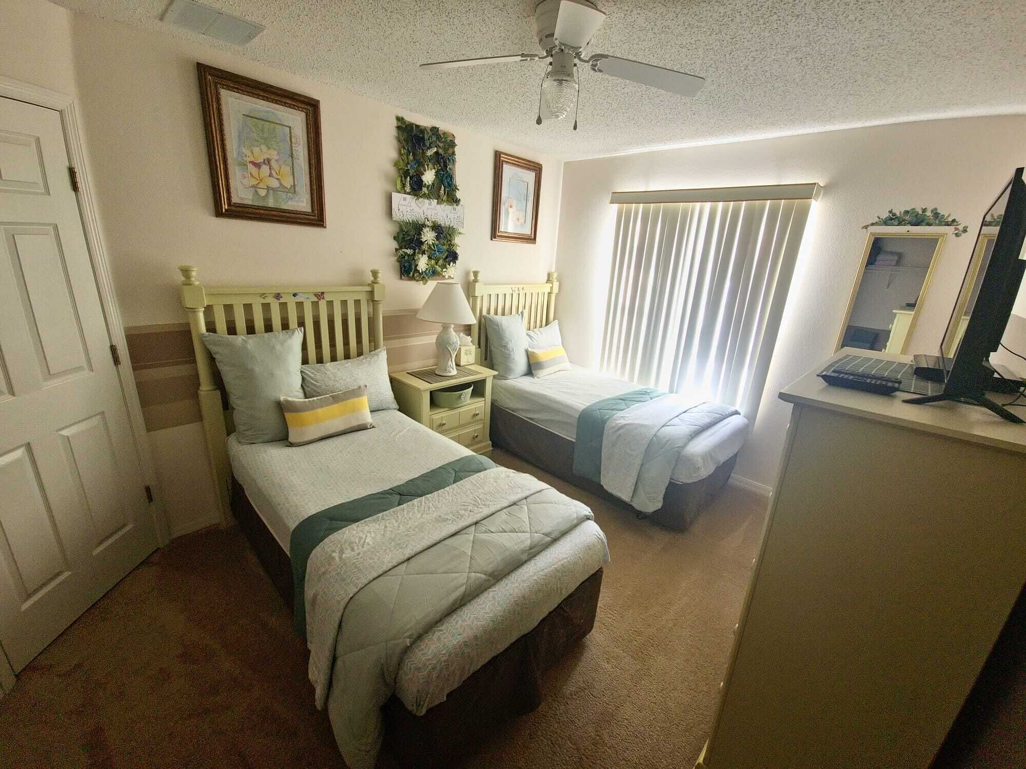 Quarto com duas camas, uma escrivaninha com computador e um ventilador de teto.