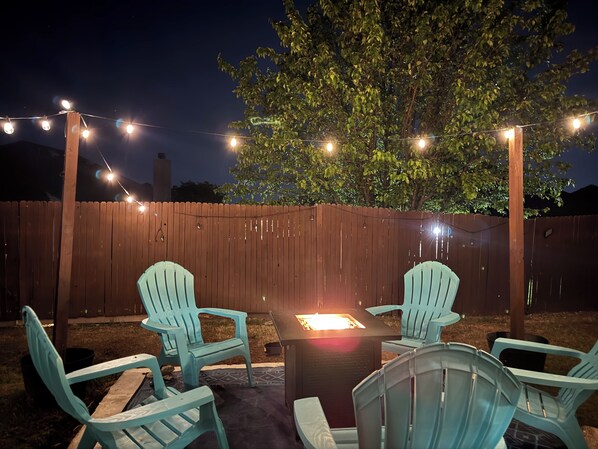 A backyard with a wooden fence, string lights, and two light blue Adirondack chairs around a square table with a fire pit.