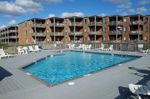 A swimming pool with lounge chairs and a multi-story building with balconies in the background. - D116A- Oceanside - Duck - Saturday Check In! (Duck)