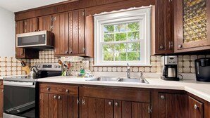 A kitchen with wooden cabinets, a microwave, a stove, a sink, and a window with a view of trees.