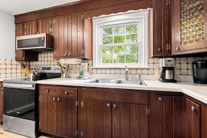 A kitchen with wooden cabinets, a microwave, a stove, a sink, and a window with a view of trees. - Minutes to Bayside Beaches, Dogs Welcome! (Eastham)
