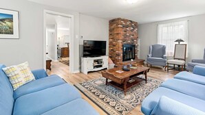 A living room with a brick fireplace, blue sofas, a TV, and a large window with sheer curtains. - Minutes to Bayside Beaches, Dogs Welcome! (Eastham)