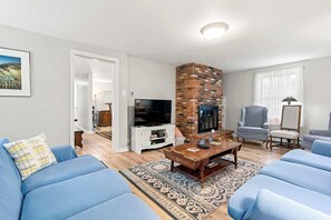 A living room with a brick fireplace, blue sofas, a TV, and a large window with sheer curtains. - Minutes to Bayside Beaches, Dogs Welcome! (Eastham)