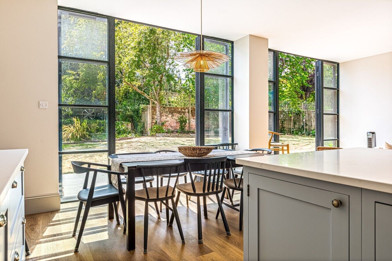 A modern kitchen with large windows, a dining area with chairs and a table, and a hanging light fixture.
