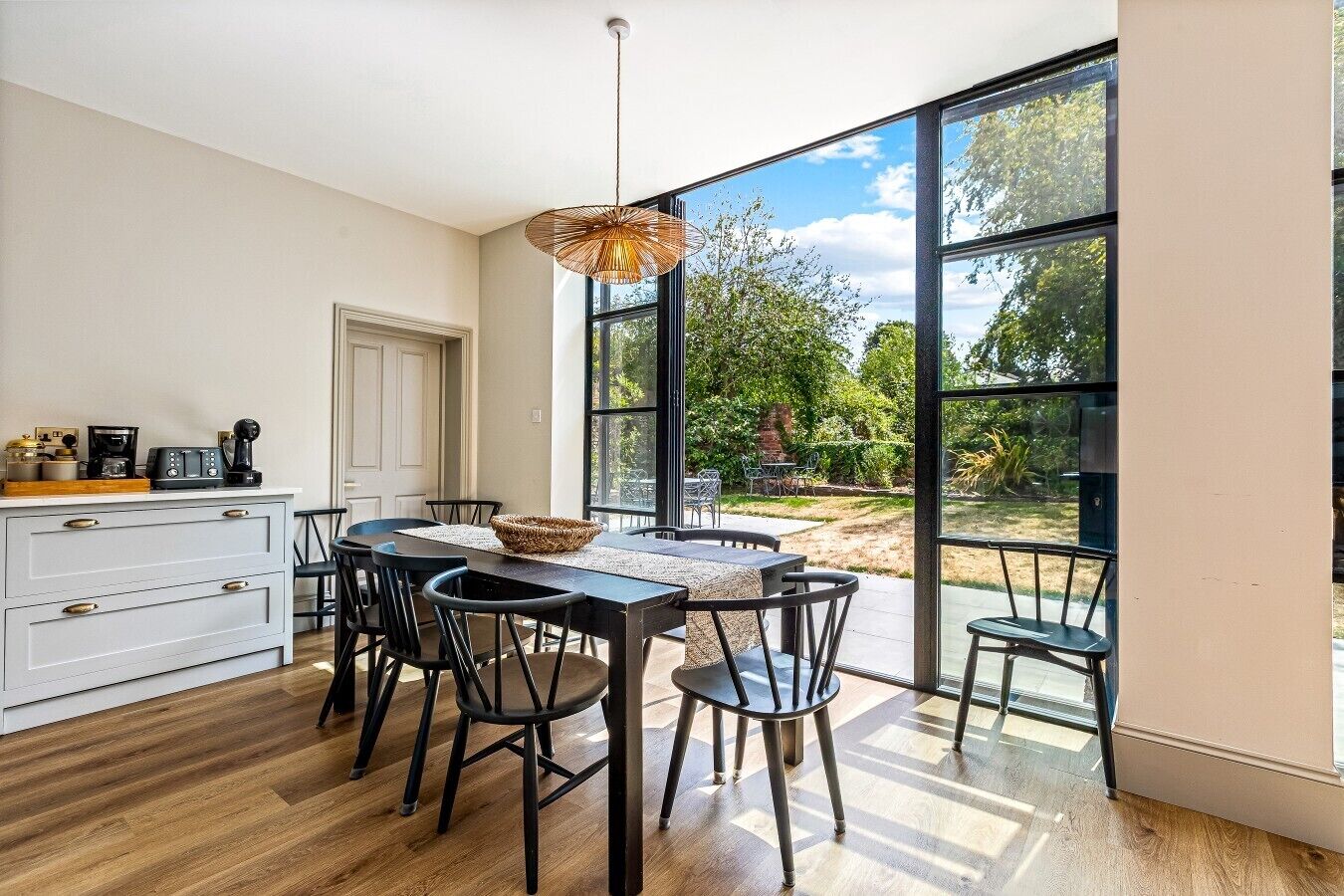A dining area with a table and chairs, a coffee machine, and a view of the outdoors through large windows.