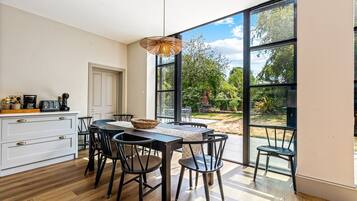 A dining area with a table and chairs, a coffee machine, and a view of the outdoors through large windows.