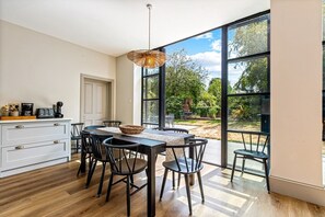 A dining area with a table and chairs, a coffee machine, and a view of the outdoors through large windows. - Montpellier House, Centrally Located, Pet Friendly (Henley-On-Thames)