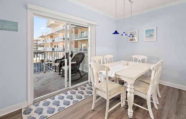 A room with a white dining table, white chairs, a wicker rocking chair, a blue patterned rug, and a view of a balcony with a railing. - Watch the Fireworks From Your Balcony!!! (Ocean City)