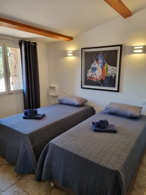 Ein Zimmer mit zwei Betten, jeweils mit Kissen und einer zusammengeklappten Decke. An der Wand hängt ein Gemälde und ein Fenster mit Vorhängen.