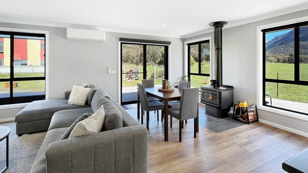 Una sala de estar moderna con sofá, mesa de comedor, sillas y estufa de leña. Amplias ventanas ofrecen vistas a un paisaje verde y un edificio al fondo.