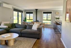 A modern living room with a gray sectional sofa, a round wooden coffee table, and a kitchen area with a dining table and bar stools.