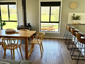 A dining area with a wooden table and chairs, a kitchen counter with stools, and large windows with a view of a grassy field.