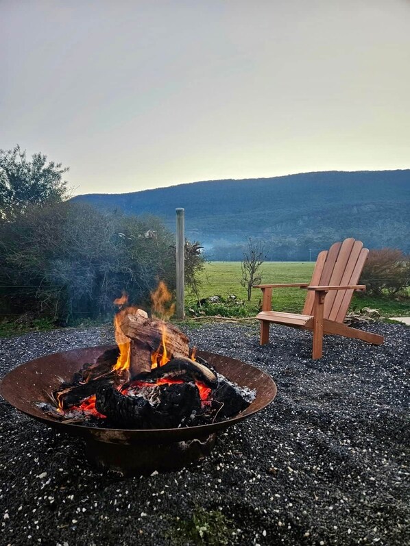 A fire pit with burning logs, a wooden chair, and a view of a hilly landscape.