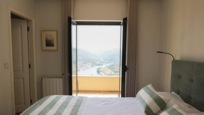 A hotel room with a bed, a view of a river and mountains, and a door to the left.