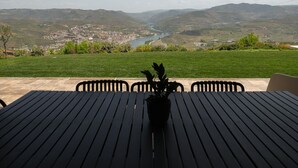 A table with chairs and a potted plant in the foreground, overlooking a scenic landscape with a river and hills.
