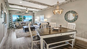 Un vaste salon doté d’une grande table à manger, de chaises blanches et d’un lustre. De grandes fenêtres offrent une vue sur l’océan.