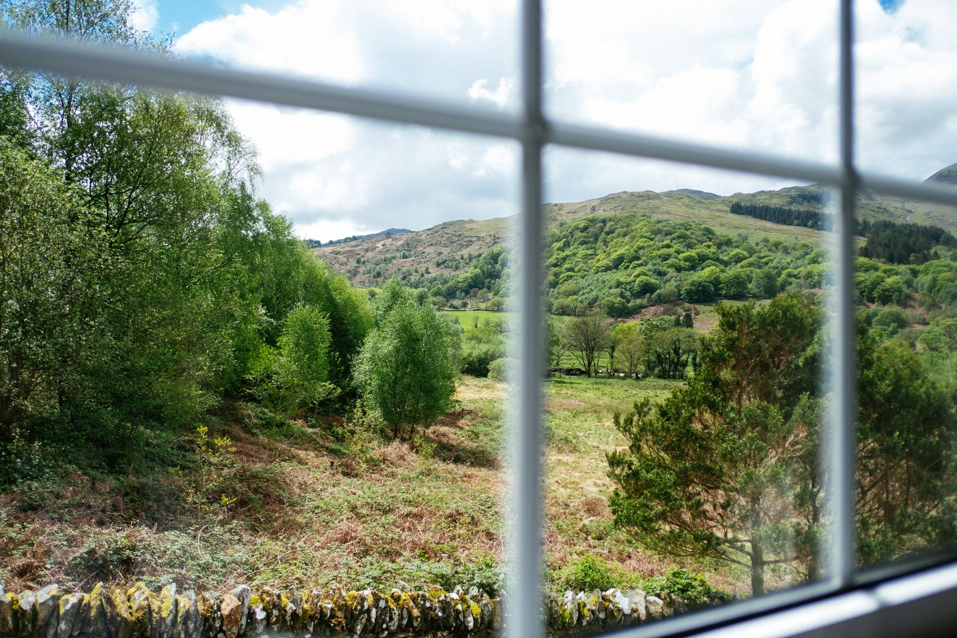 A view through a window of a rural landscape with hills, trees, and a stone wall.
