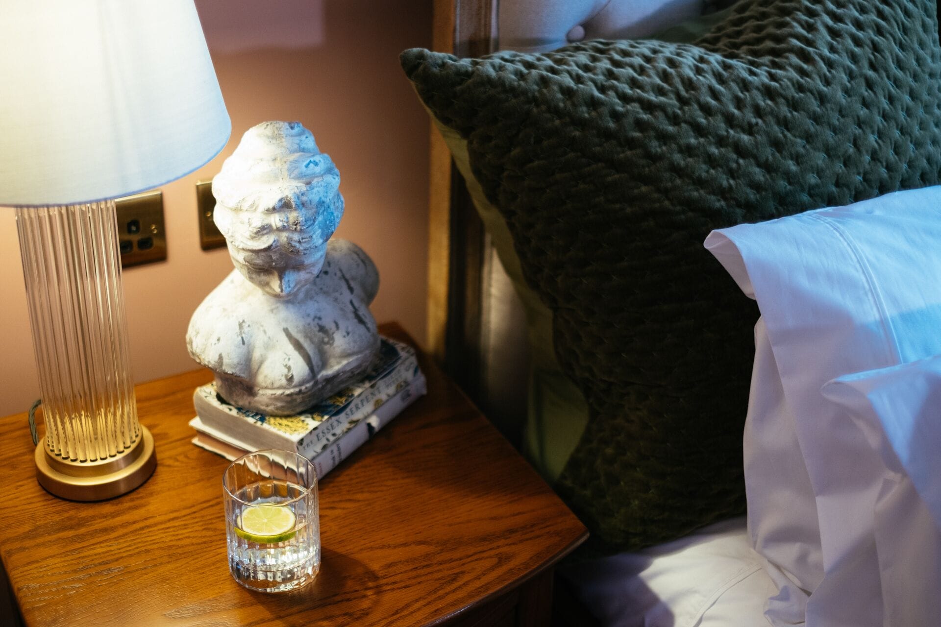 A statue on a bedside table, a glass with a lime slice, a lamp, and a green pillow.