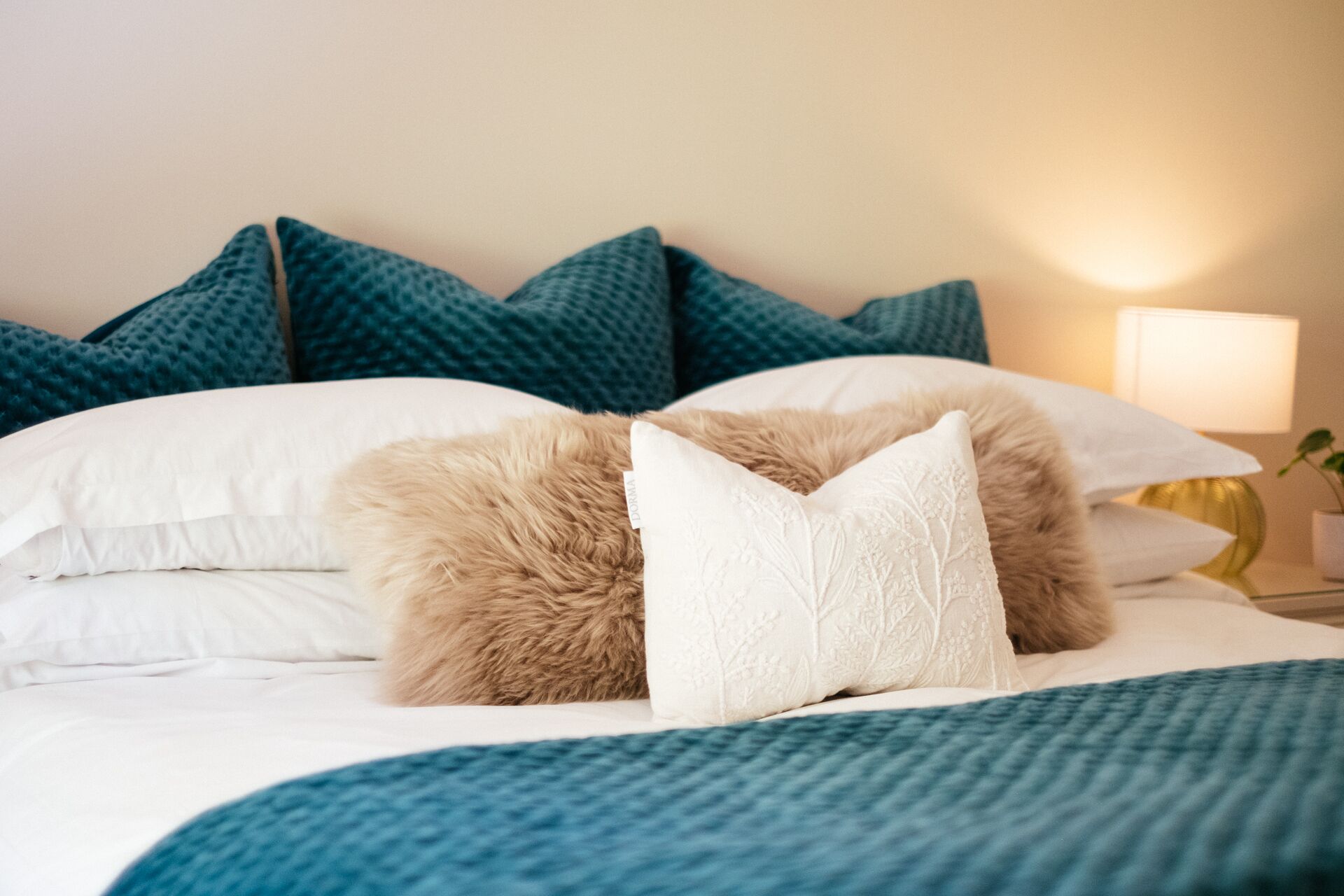 Una cama bien hecha con ropa de cama blanca y azul, un almohadón marrón mullido y almohadillas color turquesa en la cabecera.