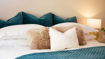 Una cama bien hecha con ropa de cama blanca y azul, un almohadón marrón mullido y almohadillas color turquesa en la cabecera.