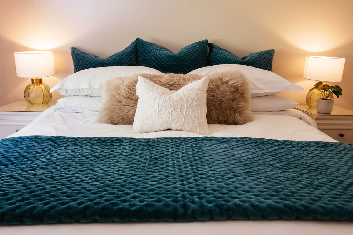 A neatly made bed with a teal duvet, white pillows, and a decorative pillow with a tree design. There are two bedside lamps with gold bases and white shades.