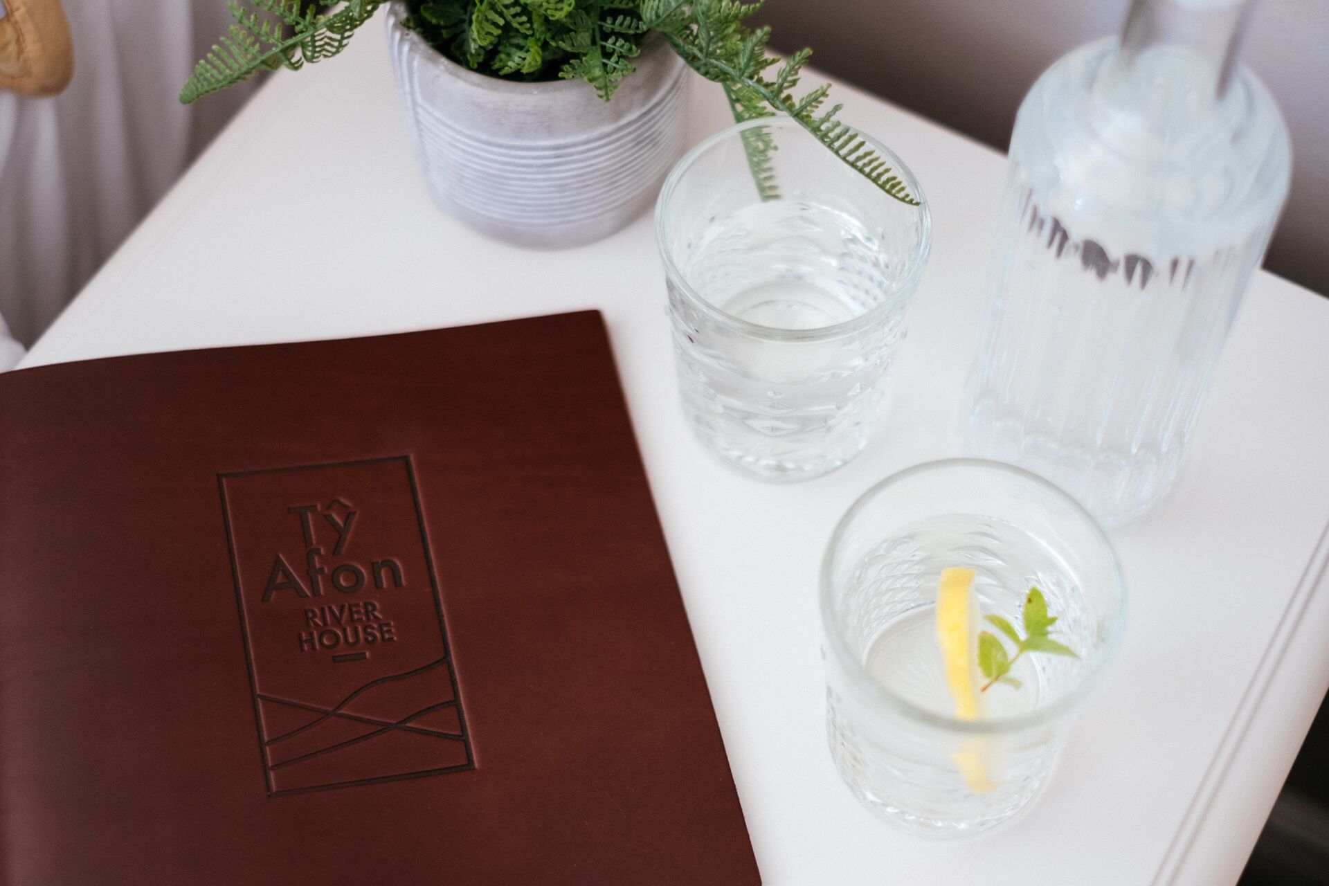 Una carpeta de cuero marrón con texto en relieve, un vaso de agua con una rodaja de limón, un vaso de agua y una botella de agua sobre una mesa blanca.