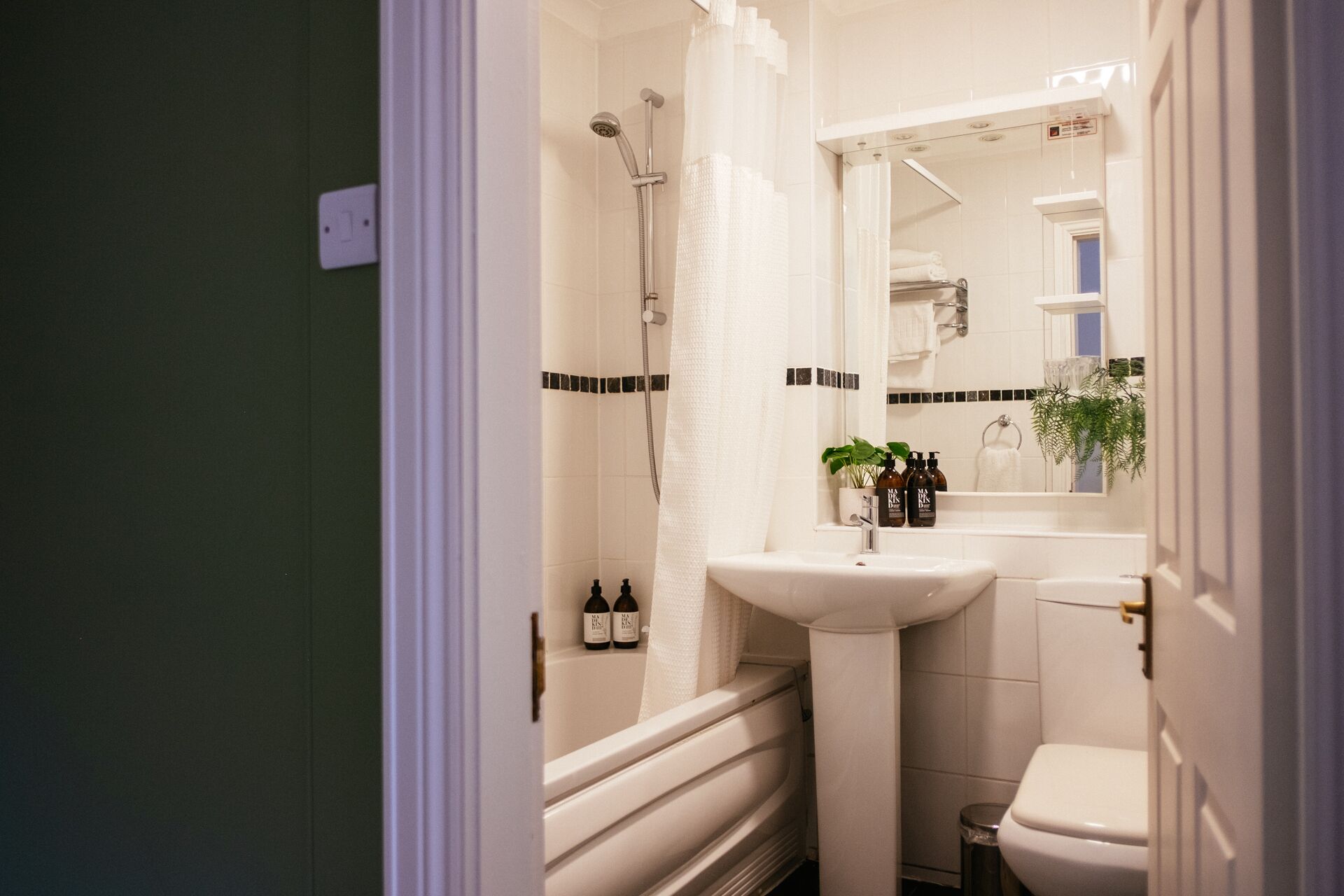 A bathroom with a white sink, a mirror, a shower, and a toilet. There are bottles of shampoo and conditioner on the sink.