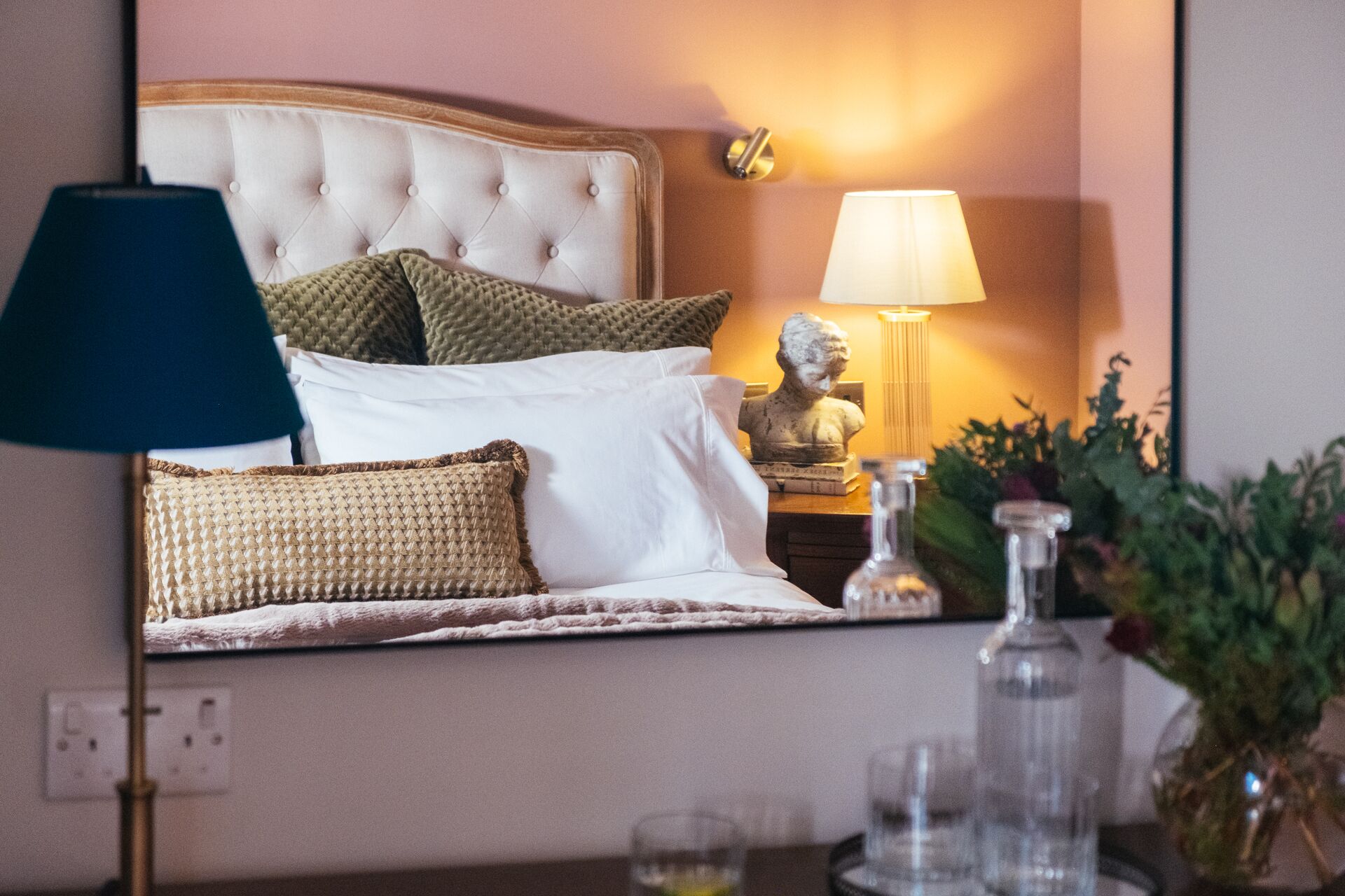 A neatly made bed with a tufted headboard, a bedside lamp, a statue, and a vase with flowers.