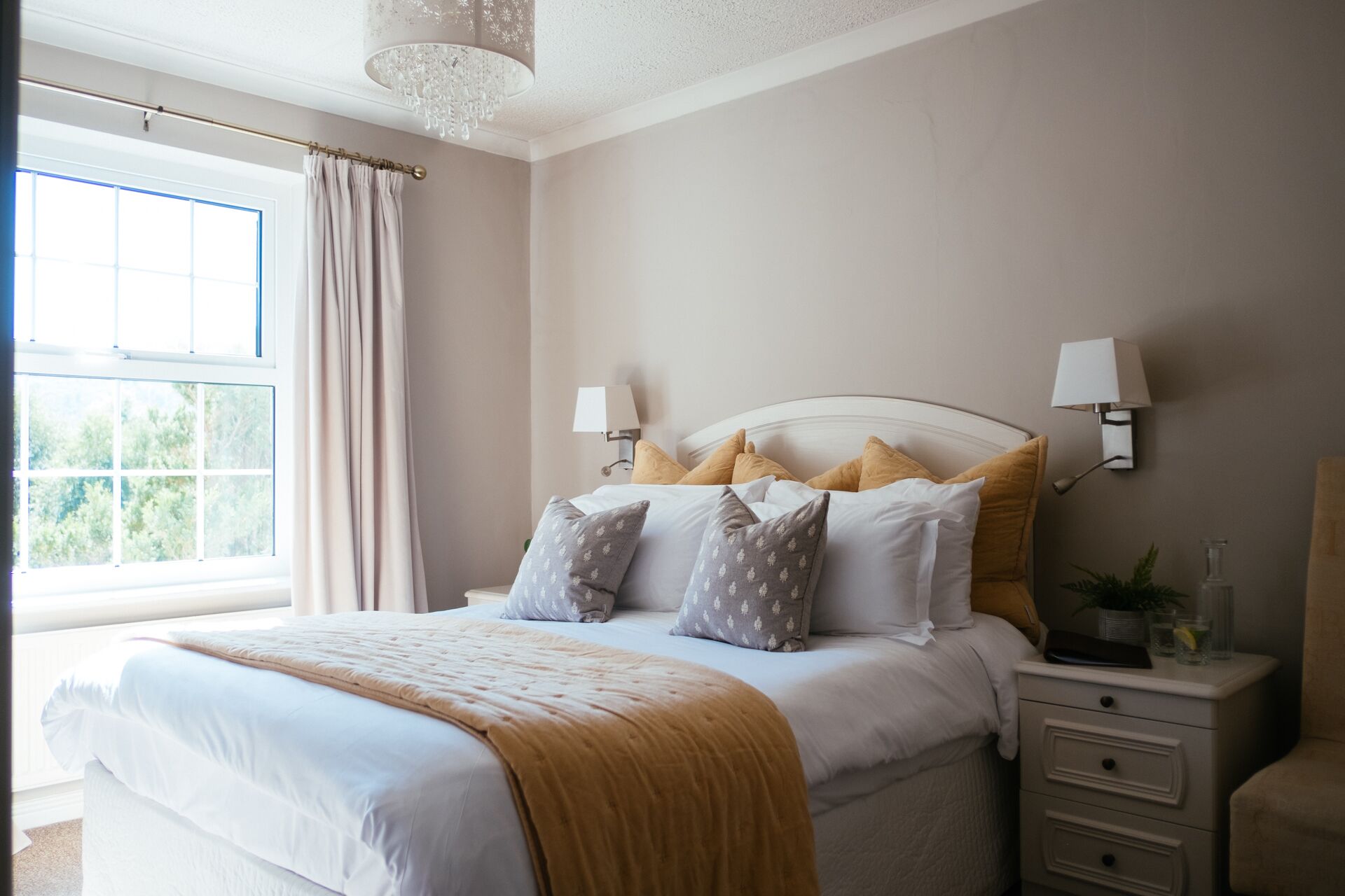 A neatly arranged bedroom with a large bed, bedside lamps, a nightstand, and a view of greenery through the window.