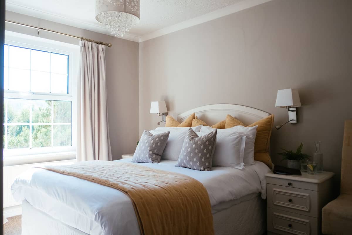 A neatly arranged bedroom with a large bed, bedside lamps, a nightstand, and a view of greenery through the window.