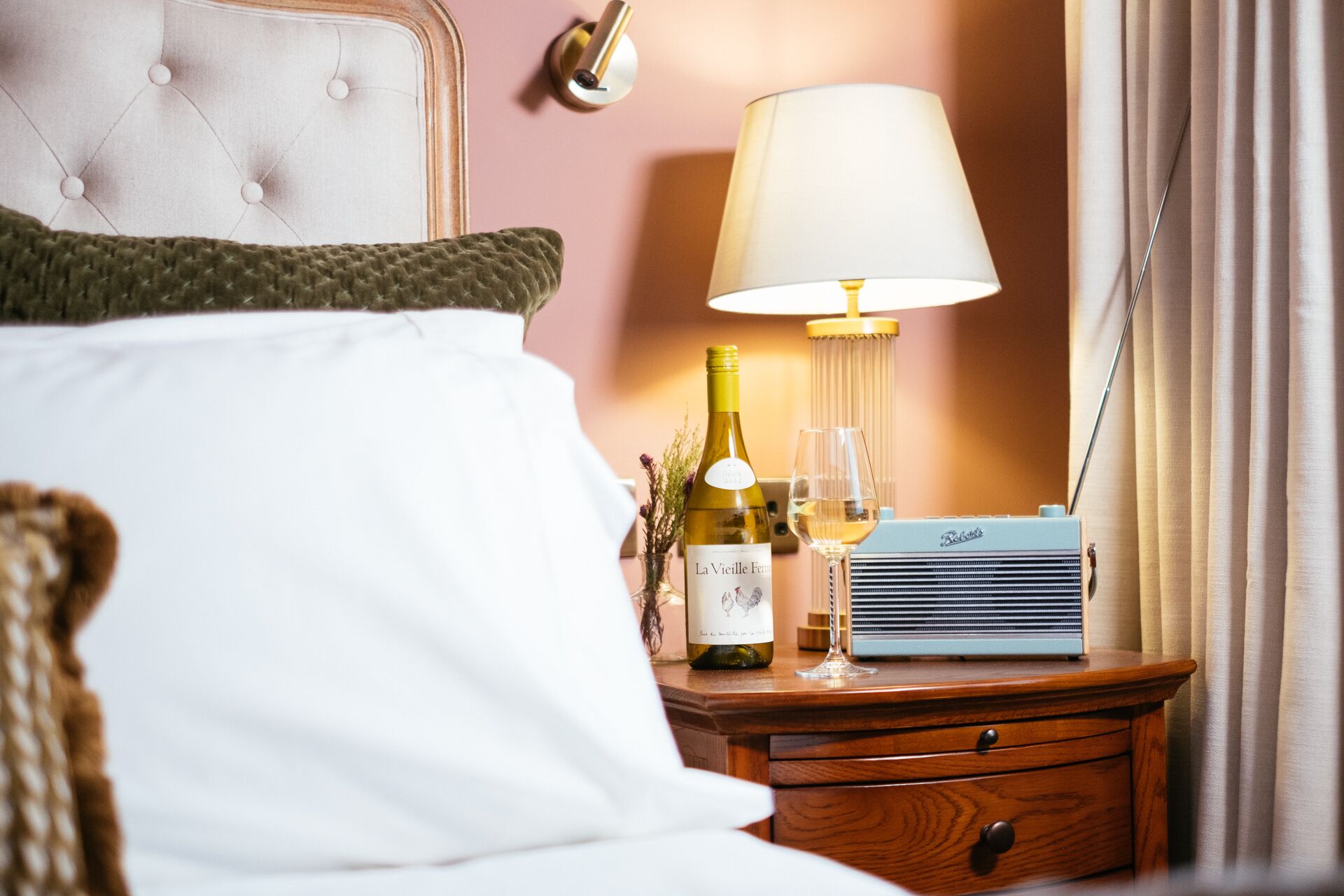 A neatly made bed with a white pillow and a green knitted throw. A bedside table holds a bottle of wine, a glass, and a vintage radio.