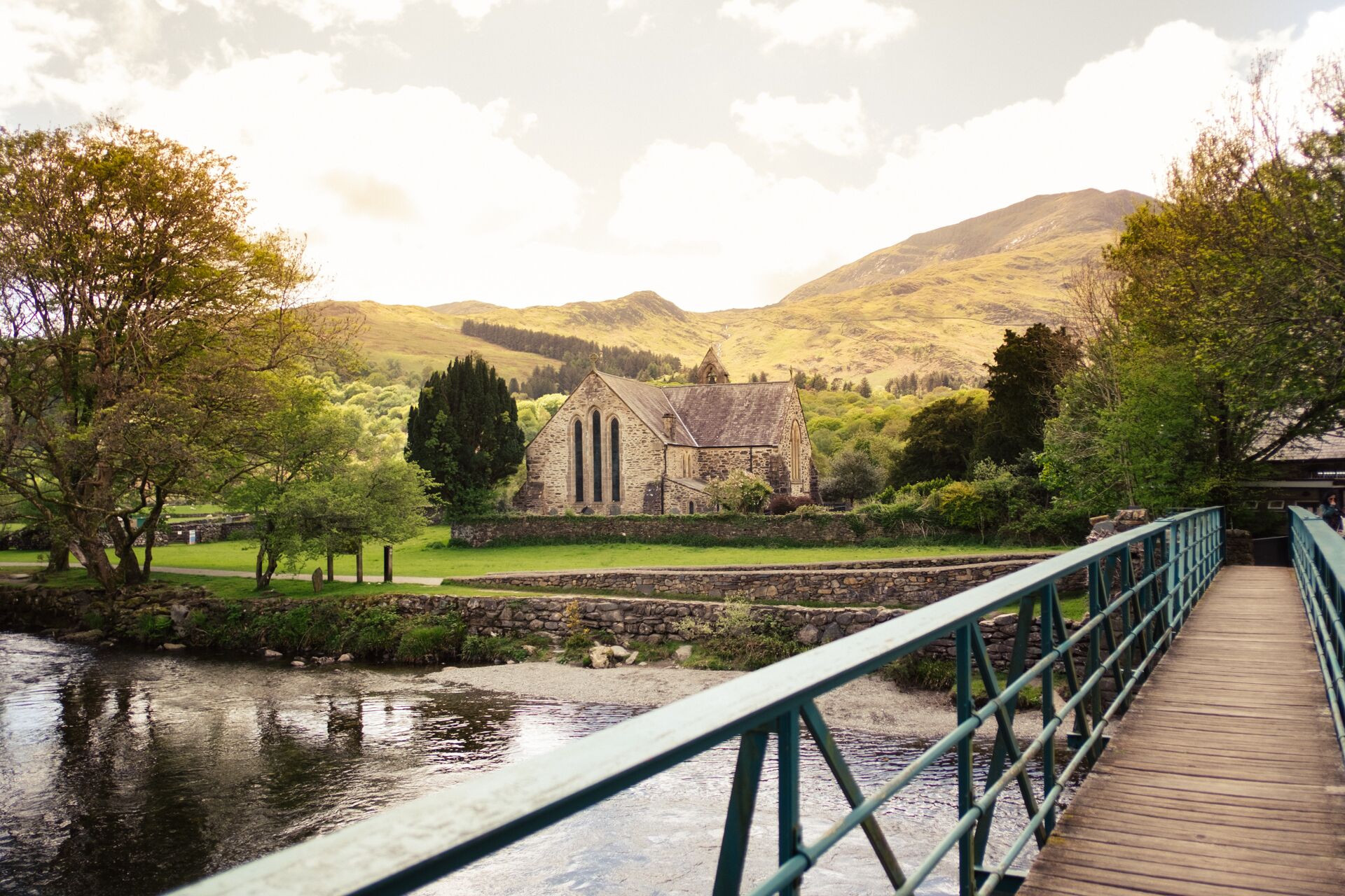 A stone church with a wooden bridge leading to it, surrounded by greenery and hills.