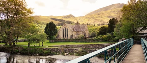 A stone church with a wooden bridge leading to it, surrounded by greenery and hills.