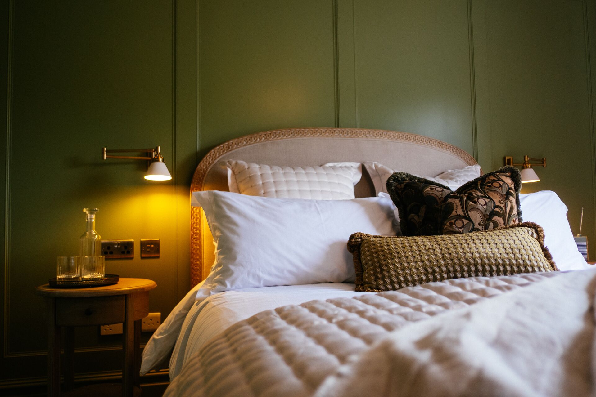Una cama bien hecha con sábanas blancas y almohadones decorativos, una mesita de noche con una lámpara y una lámpara de pared.