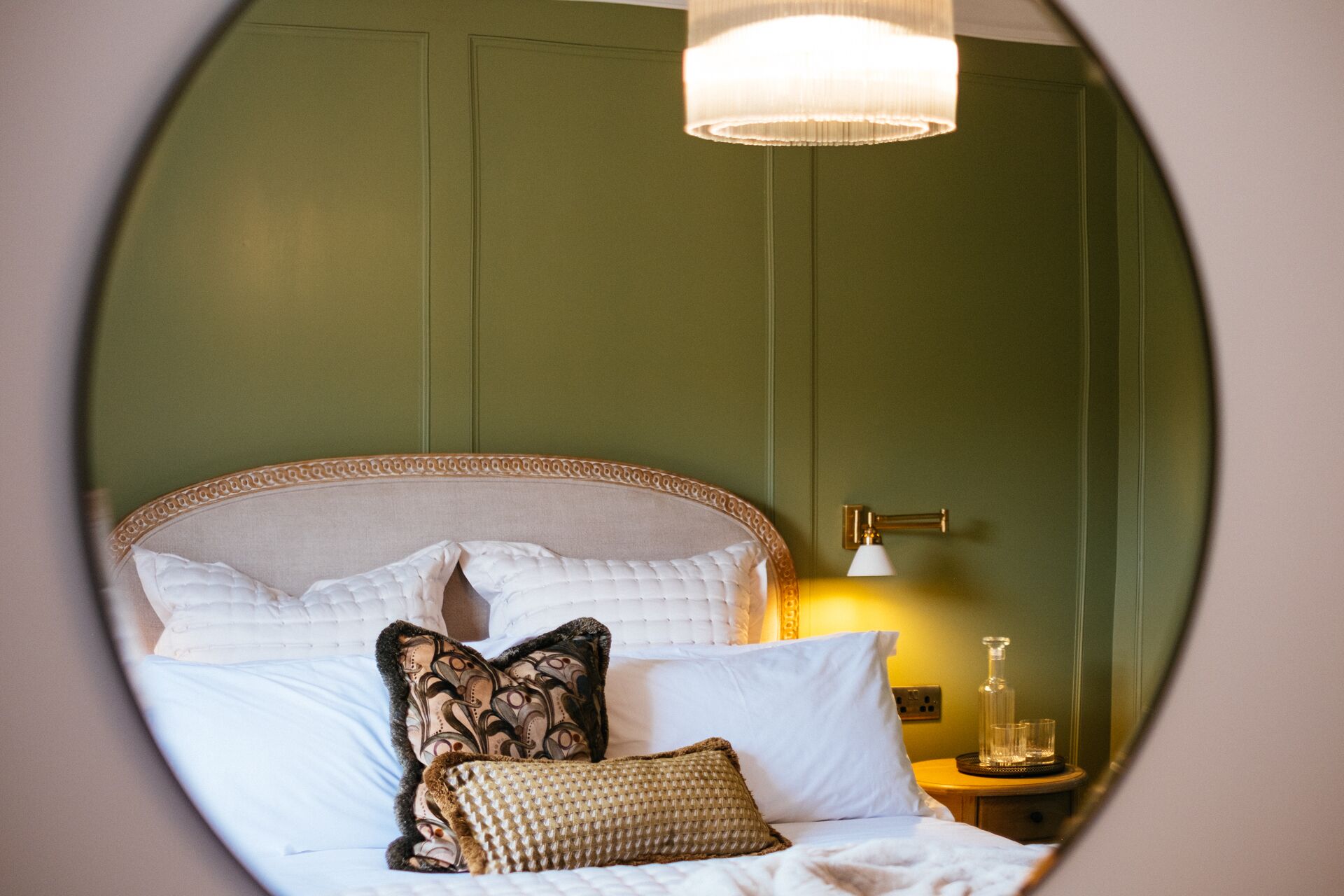 A circular view of a bed with a tufted headboard, multiple pillows, and a bedside table with a lamp and glassware.