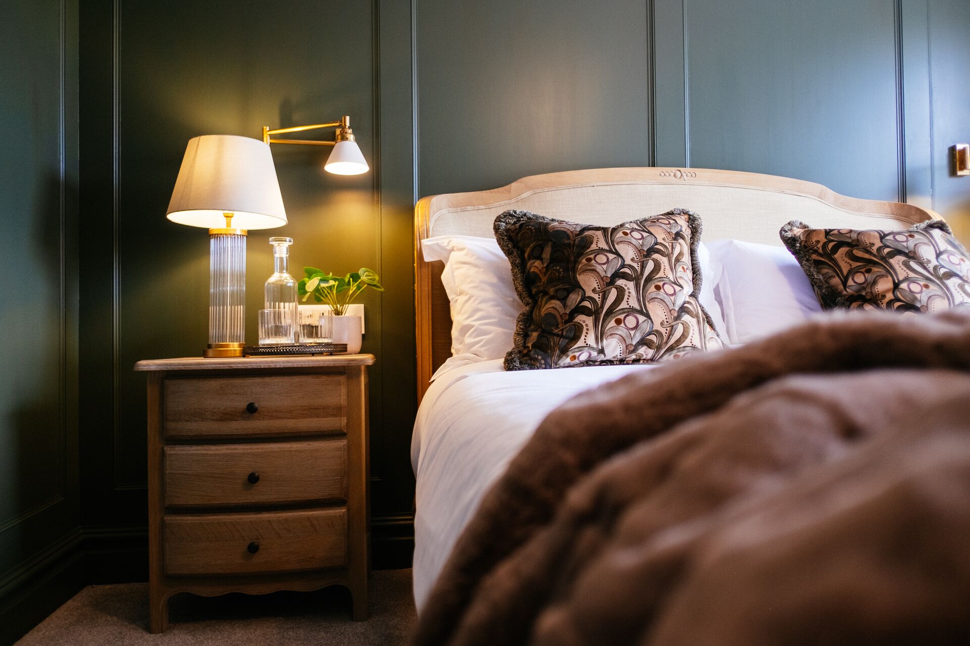Una cama bien hecha con almohadones estampados, una lámpara de noche y una pequeña mesita de madera con cajones y una botella de vidrio.