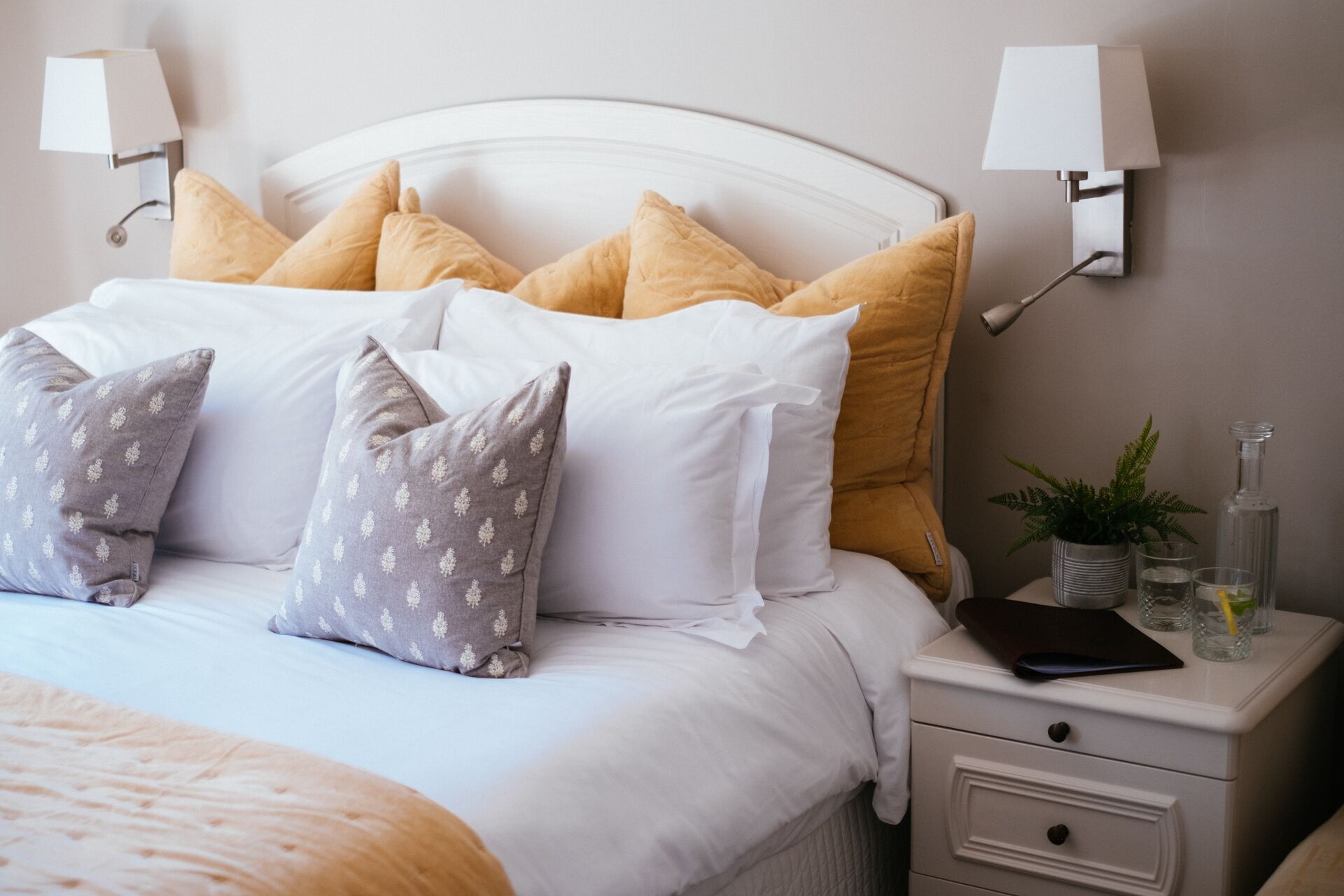Una cama bien tendida con almohadas blancas y estampadas, una mesita de noche con una lámpara, una planta y una botella.
