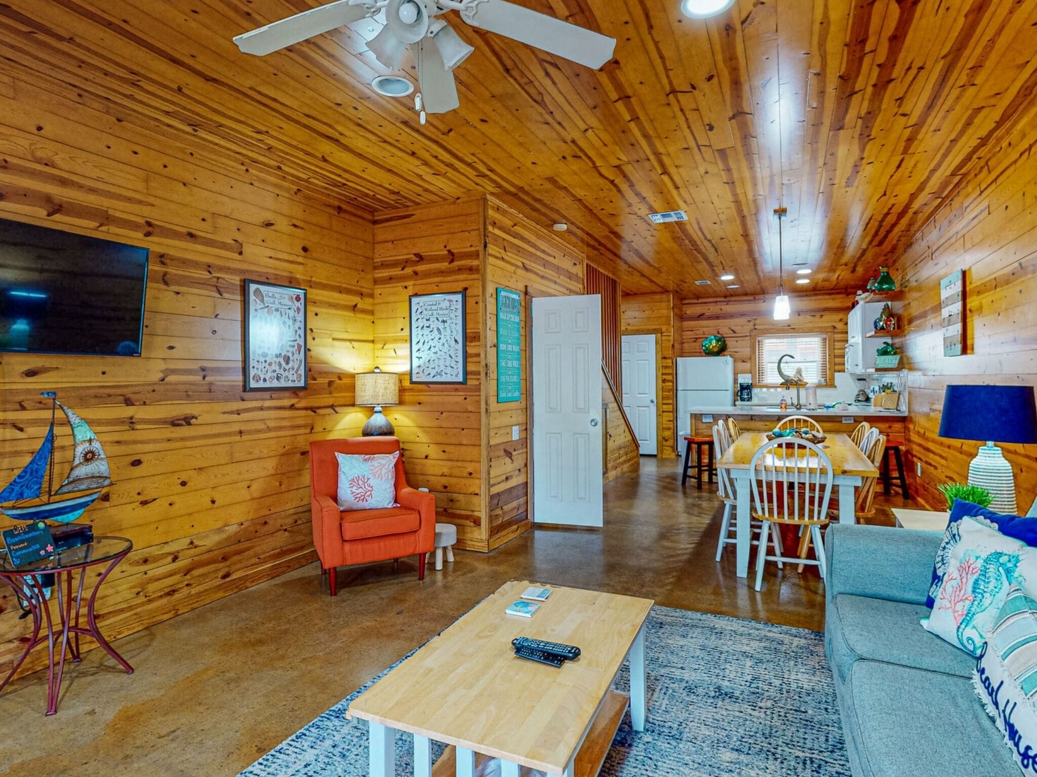 A cozy cabin interior with wooden walls, a dining area, a living room with a sofa and coffee table, and a kitchen area.
