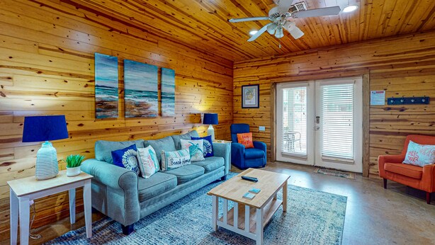 Ein gemütliches Blockhauszimmer mit Holzvertäfelung, einer grauen Couch, einem blauen Sessel, einem kleinen Holztisch und einem Deckenventilator.