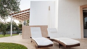 Zwei Korbsessel mit weißen Kissen auf einer Steinterrasse, darüber eine Pergola und im Hintergrund eine weiße Wand.