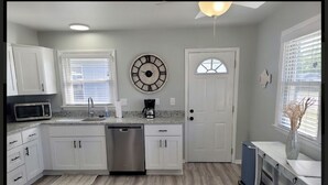 A modern kitchen with white cabinets, a stainless steel dishwasher, a microwave, and a coffee maker. There is a clock on the wall and a window with blinds. - Renovated 2 Bedroom, near VB Vibe district, Surf Park & Dome! (Virginia Beach)