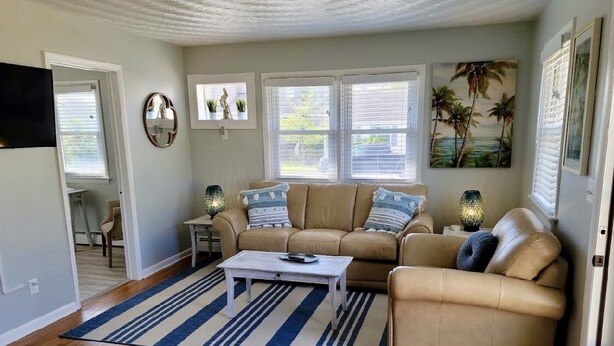 A living room with a beige sofa, a coffee table, and a striped rug. There are windows with blinds, a TV mounted on the wall, and a painting of palm trees. - Renovated 2 Bedroom, near VB Vibe district, Surf Park & Dome! (Virginia Beach)