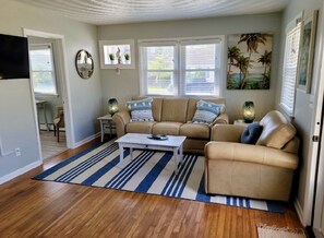 A living room with a beige sofa, a coffee table, and a striped rug. There are windows with blinds, a TV mounted on the wall, and a painting of palm trees.