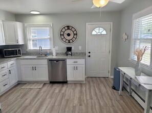 A modern kitchen with white cabinets, a stainless steel dishwasher, a microwave, and a coffee maker. There is a clock on the wall and a window with blinds.