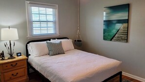 A bedroom with a bed, bedside table, lamp, window, and artwork. - Renovated 2 Bedroom, near VB Vibe district, Surf Park & Dome! (Virginia Beach)