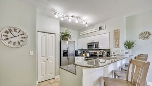 Una cocina con un reloj, un refrigerador, un microondas, un lavabo y una zona de comedor con sillas.