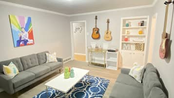 A modern living room with a grey sofa, a white coffee table, and a blue patterned rug. There are two guitars on the wall and a bookshelf with various items.